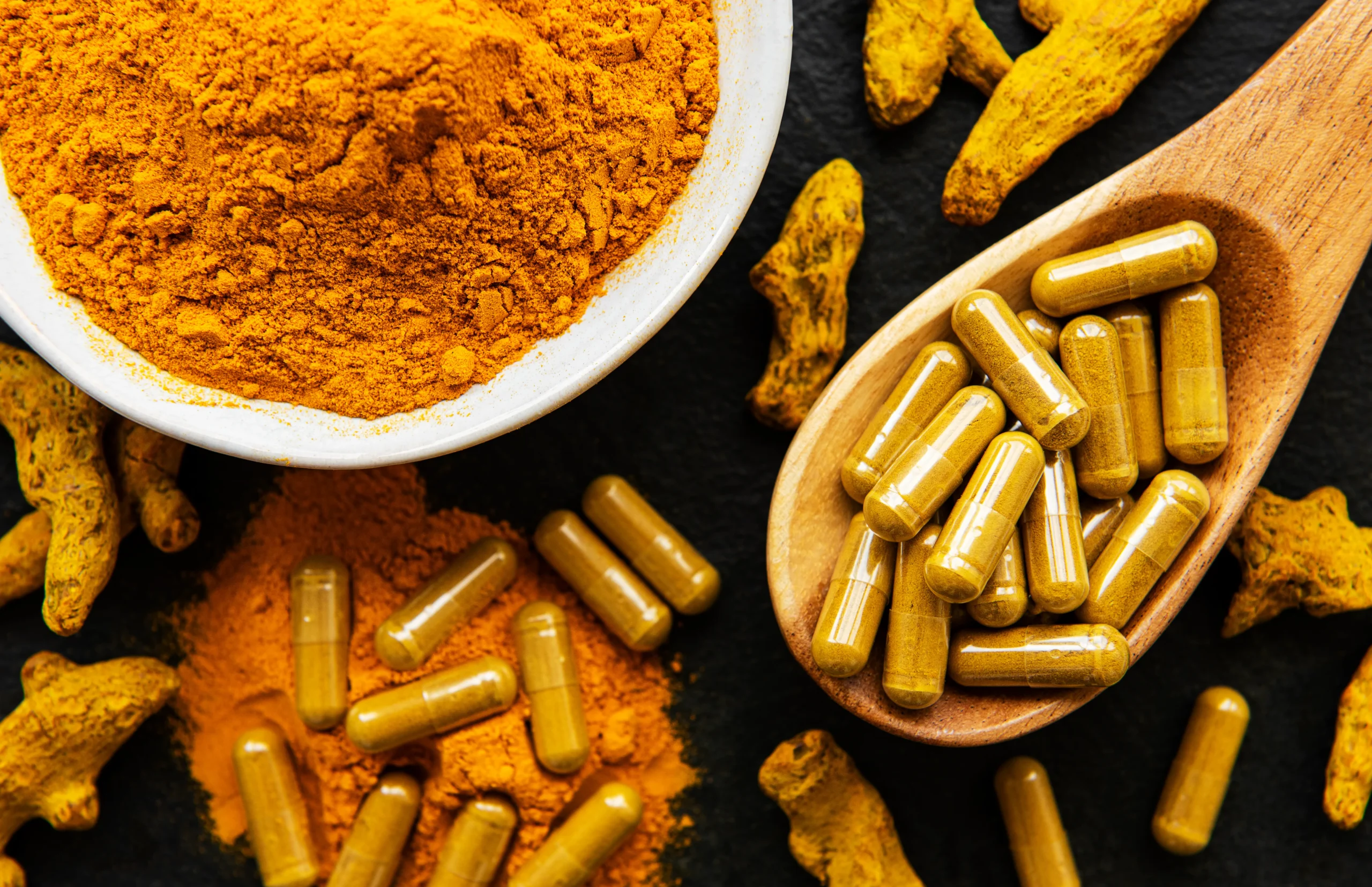 Turmeric powder, dried turmeric roots and supplement capsules arranged on a dark surface.